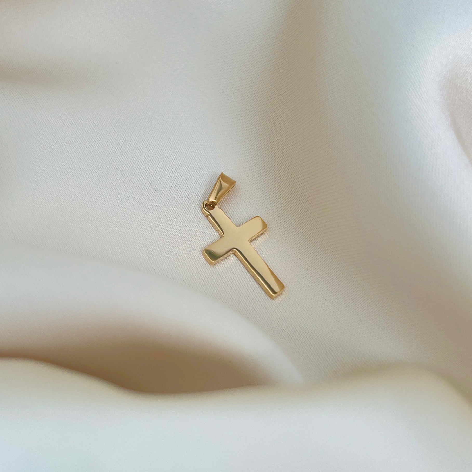 Chris Cross Pendant från Alora Avenue i närbild mot cremefärgat tyg – ett vattentåligt korshänge i 18k guldpläterat rostfritt stål. Ett allergivänligt smycke med stilren symbolik, perfekt som present till dop, konfirmation eller som tidlös accessoar i vardagen.