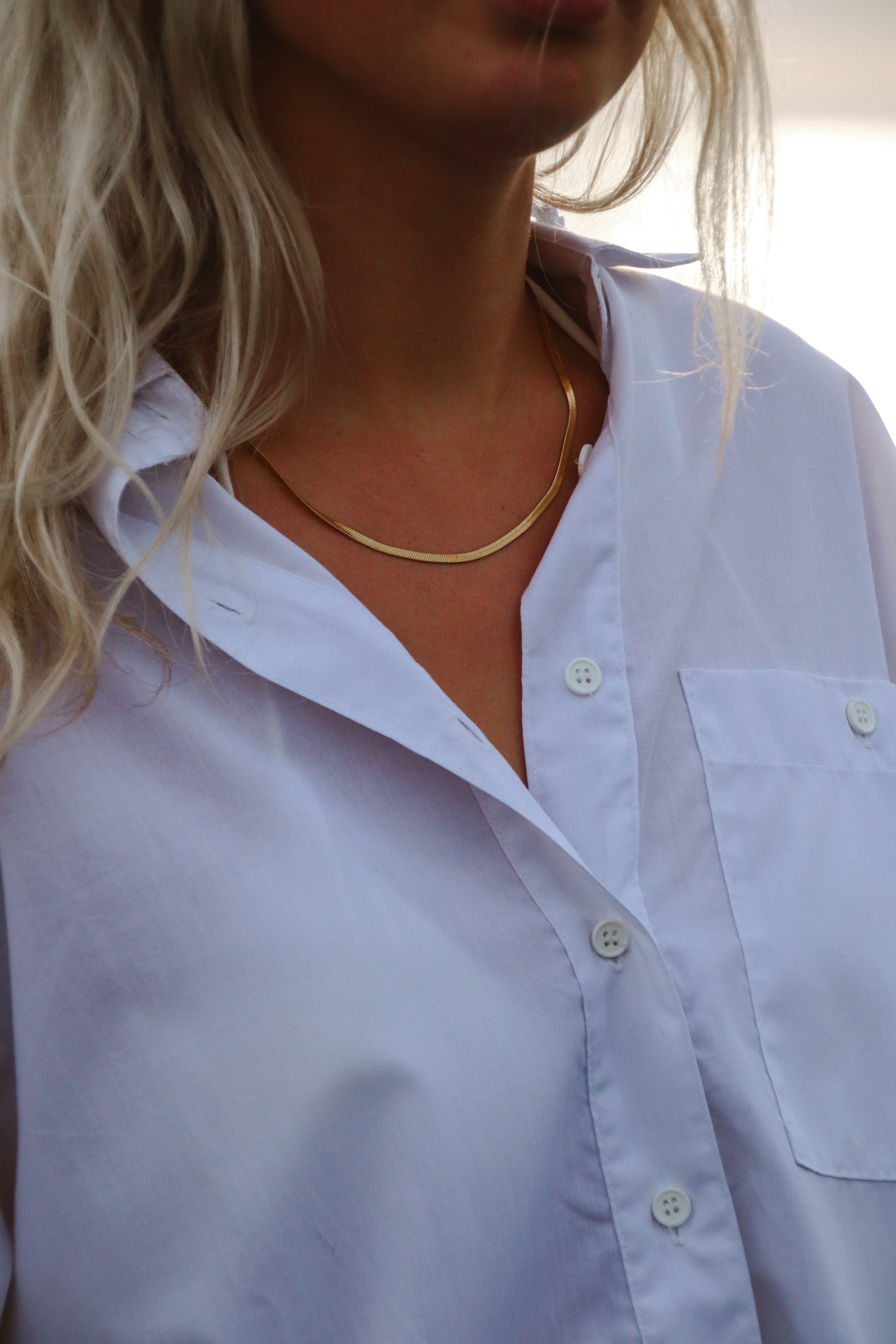 Clara Cobra Necklace – ett glansigt, platt guldfärgat halsband – bärs till en vit skjorta med öppen krage. Smycket reflekterar ljuset elegant mot huden.