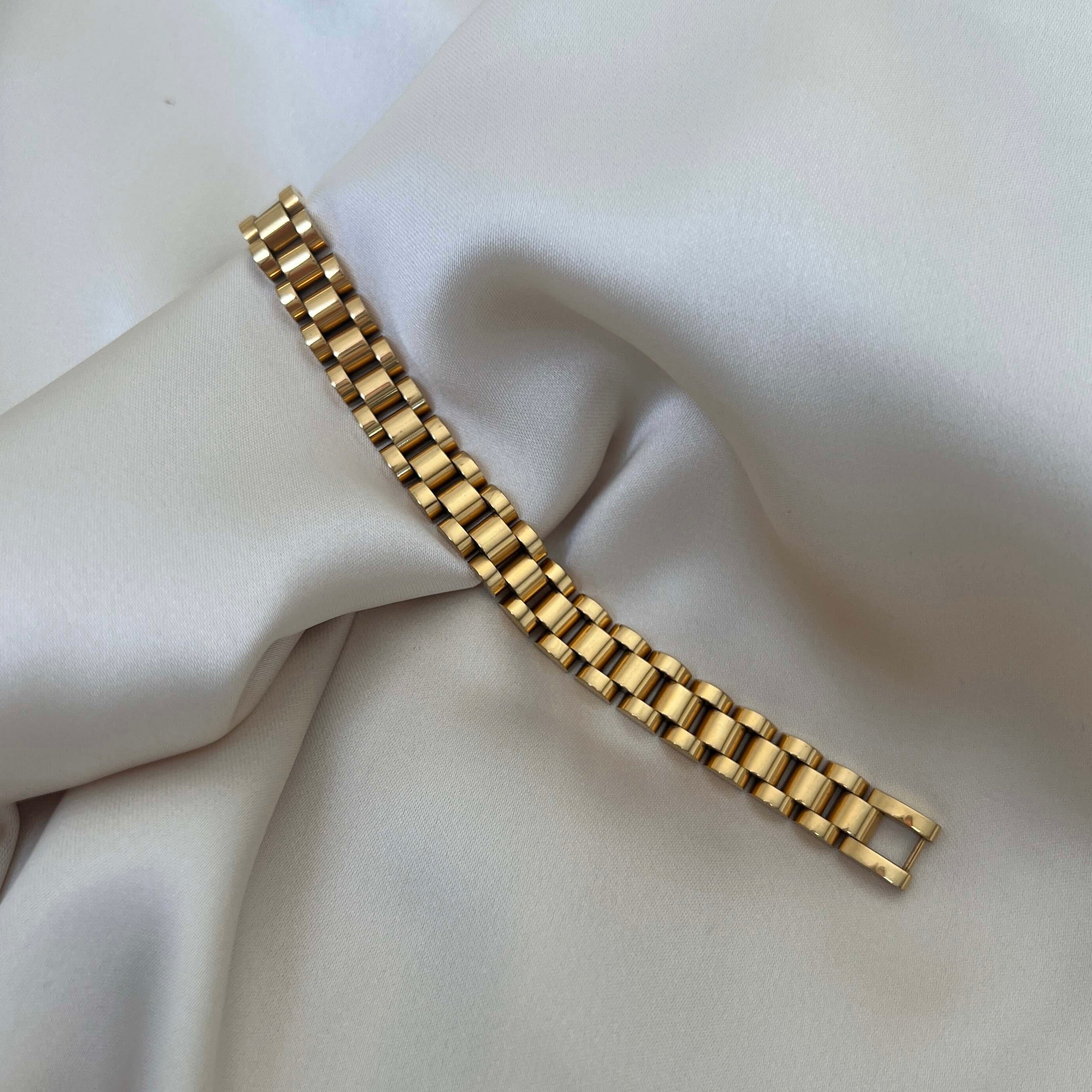 Analog Aria Wristband i guldfärgat rostfritt stål – klockinspirerat armband med tidlös design, vattentåligt och allergivänligt tack vare slitstark PVD-plätering. Ligger elegant mot en krämfärgad bakgrund.