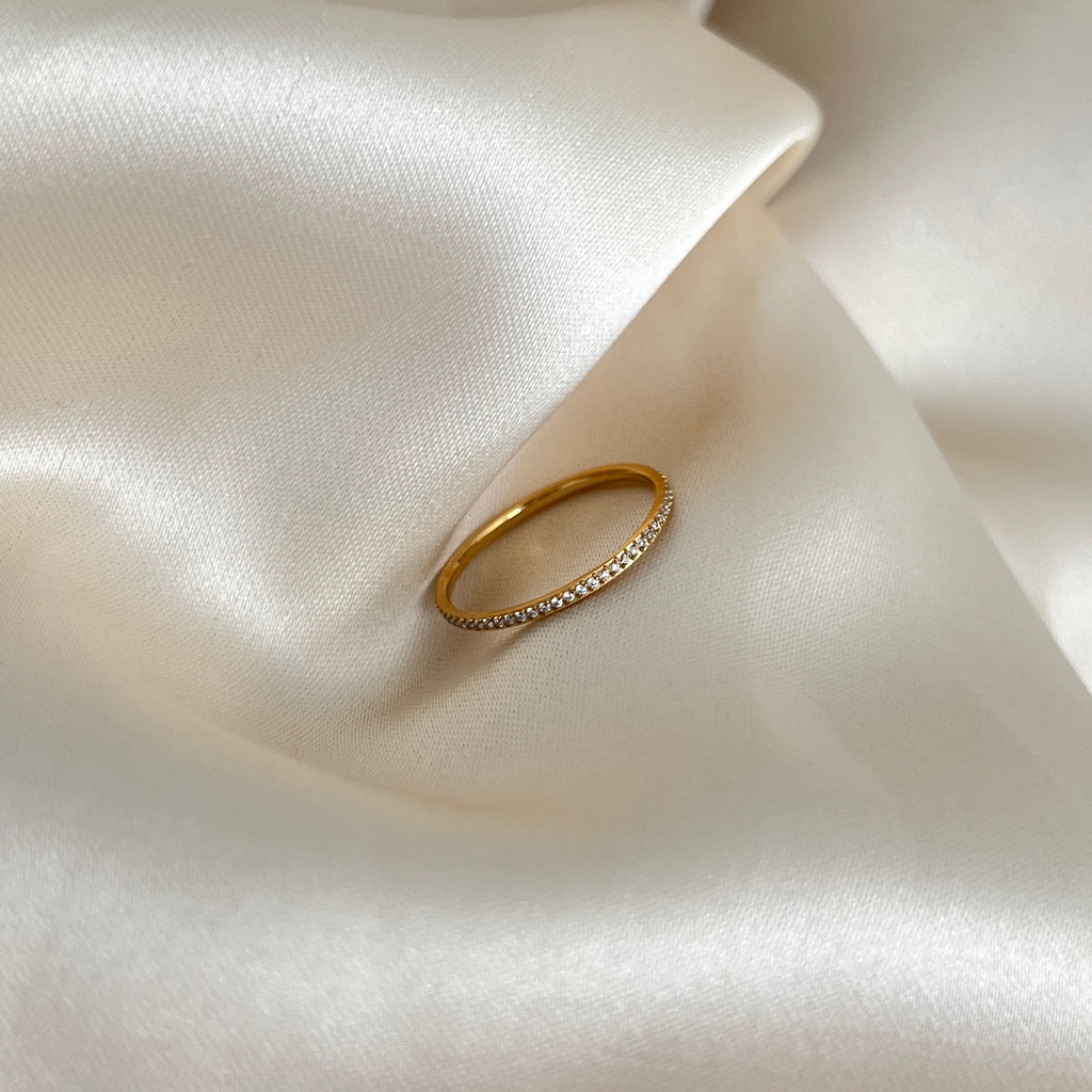 Ina Spark Ring i guld med gnistrande stenar längs nästan hela ringen, fotograferad på ljus sidenbakgrund. Tidlös, vattentålig och allergivänlig ring från Alora Avenue.