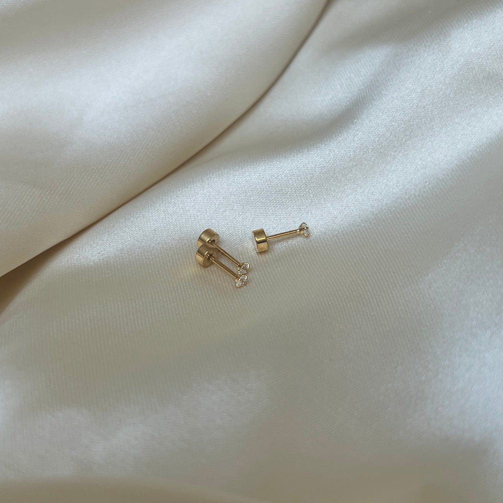 Tre Iris Star Stud-örhängen i guld med vit sten fotograferade på ljus sidenbakgrund. Tidlöst, vattentåligt och bekvämt örhänge med platt skruvbaksida från Alora Avenue.