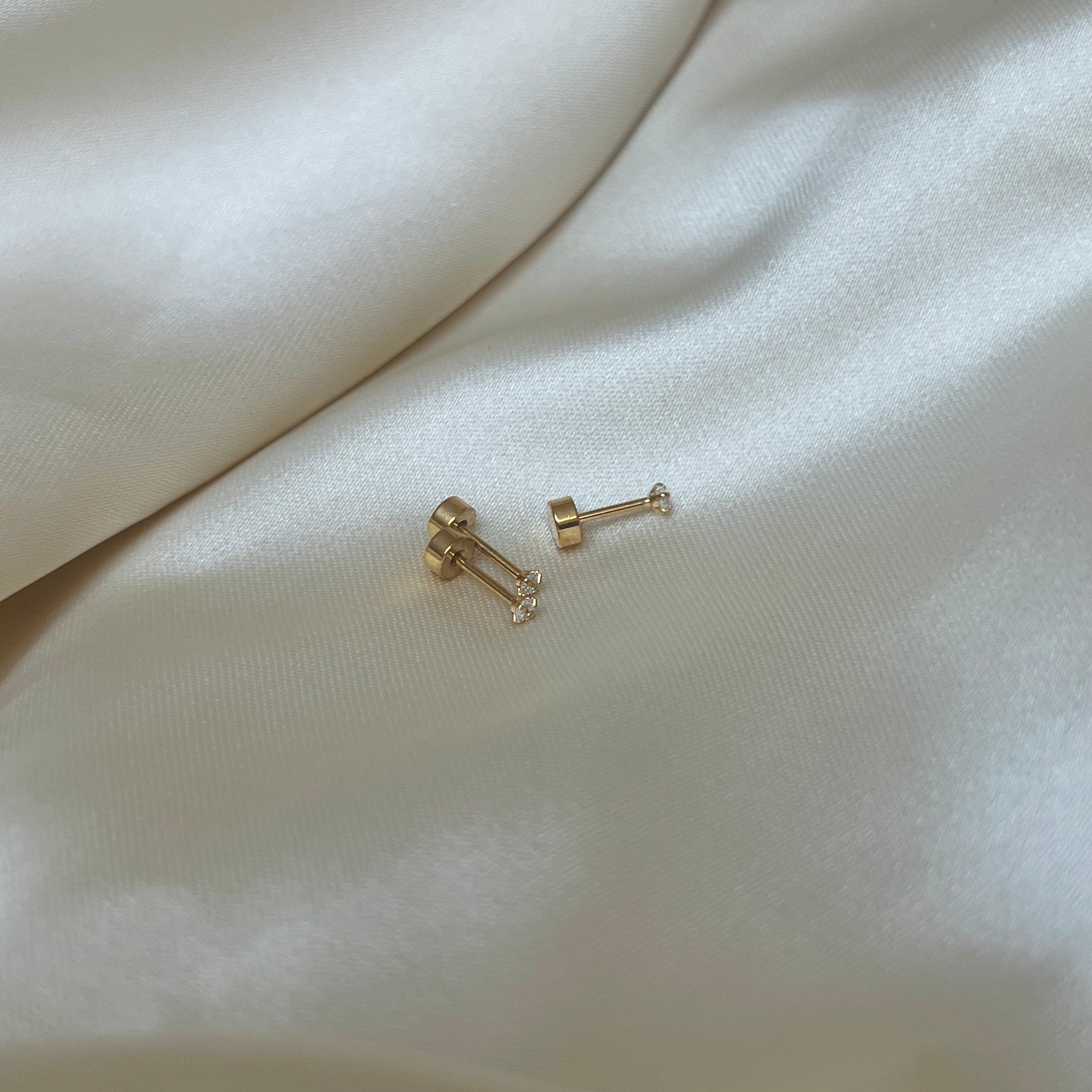 Tre Iris Star Stud-örhängen i guld med vit sten fotograferade på ljus sidenbakgrund. Tidlöst, vattentåligt och bekvämt örhänge med platt skruvbaksida från Alora Avenue.