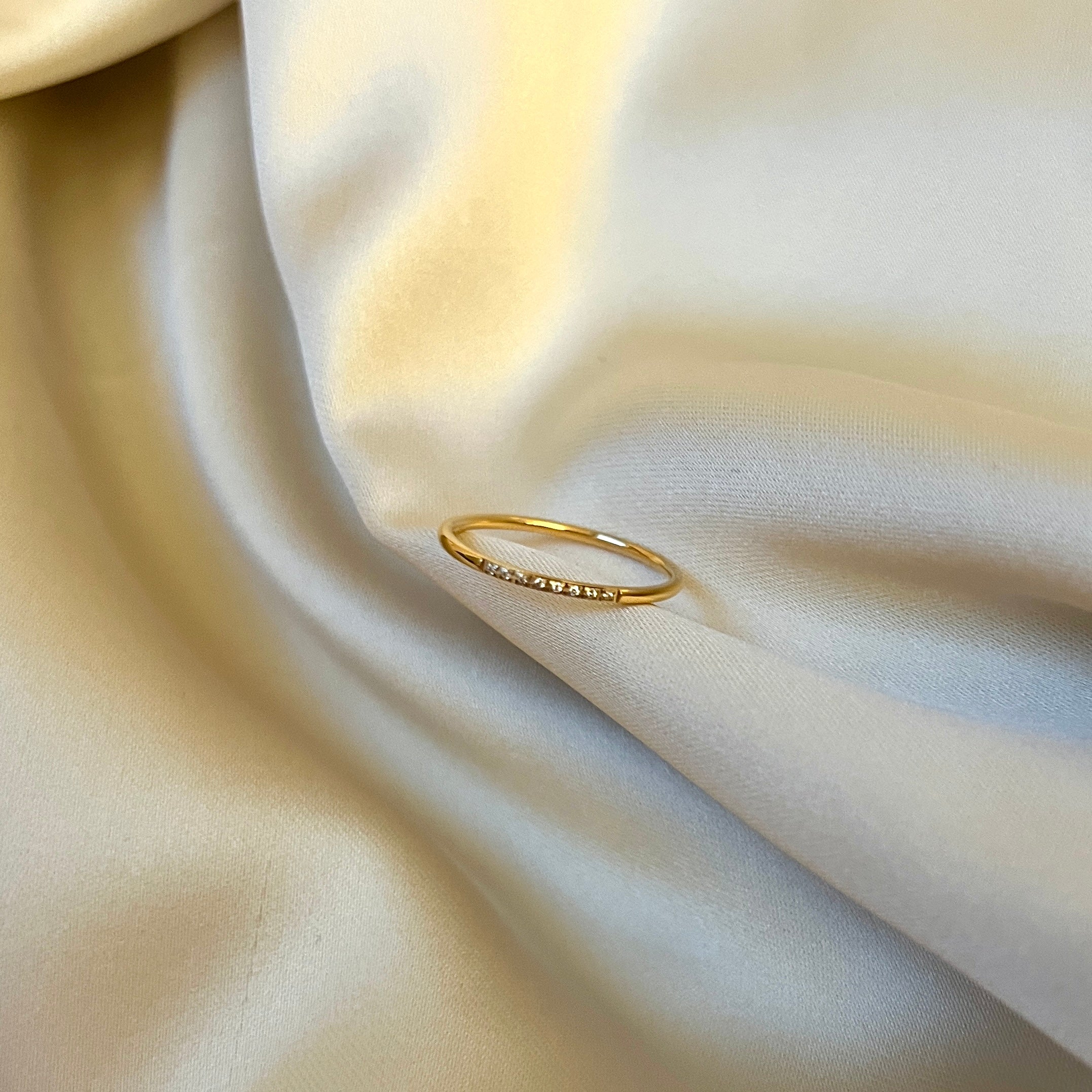 Shim Shim Ring från Alora Avenue liggande på skimrande beige tyg – en tunn guldfärgad ring med små klara kristaller i rad. Elegant, minimalistisk och vattentålig design för vardag och fest.