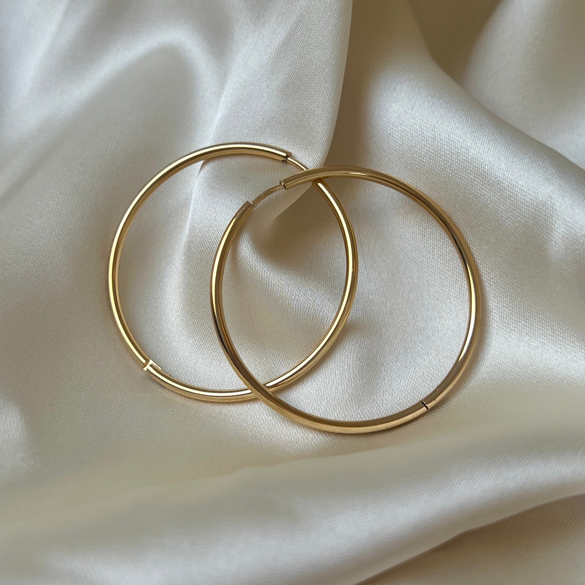 Sky Saturn Hoops i guld fotograferade mot ett ljust silkestyg – eleganta, vattentåliga och allergivänliga hoops i rostfritt stål med lyxig glans och tidlös design.