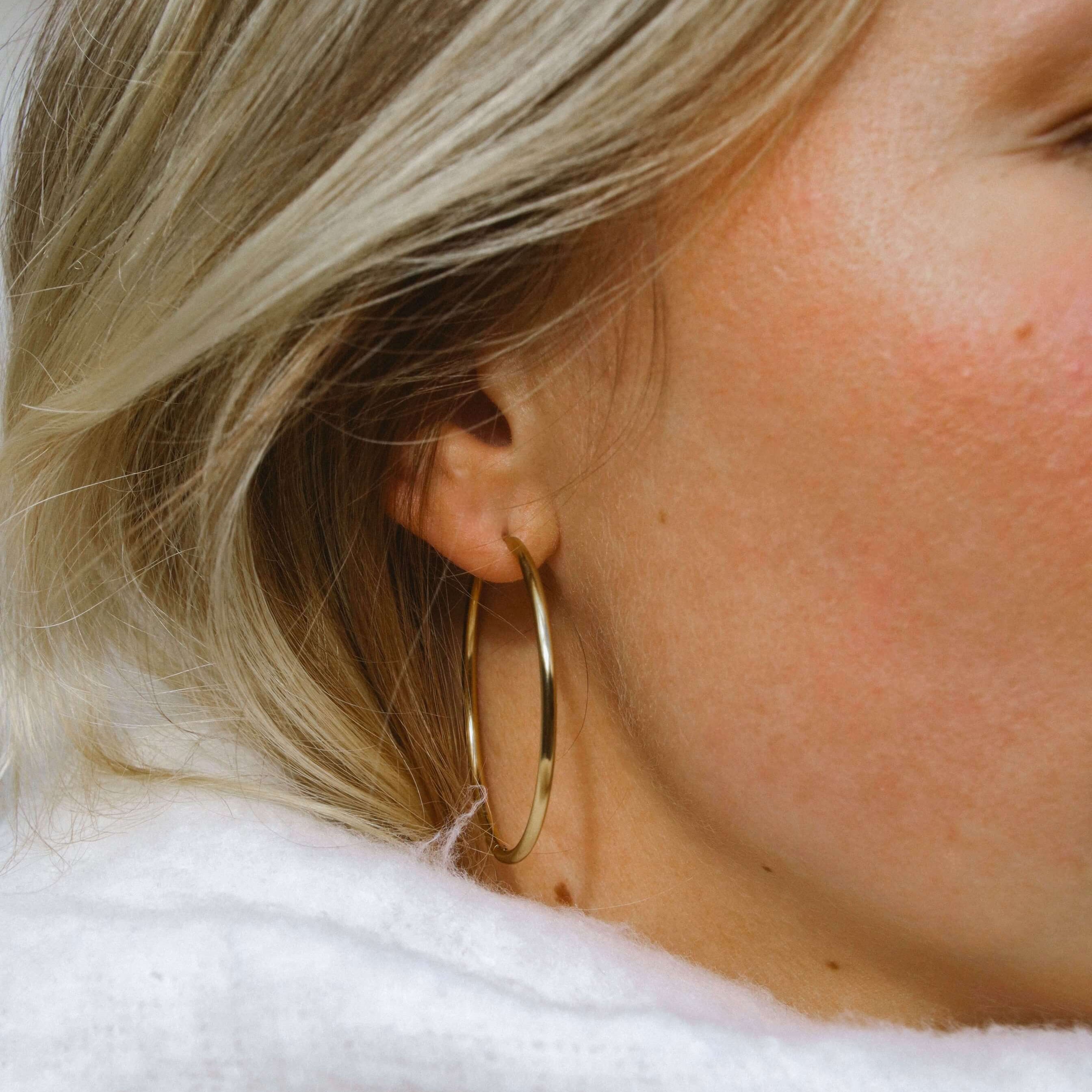 Kvinna bär Sky Saturn Hoops tillsammans med en vit halsduk – tunna guldiga hoops i rostfritt stål med PVD-plätering. Stilrena, vattentåliga och allergivänliga örhängen som ger en elegant höstlook.